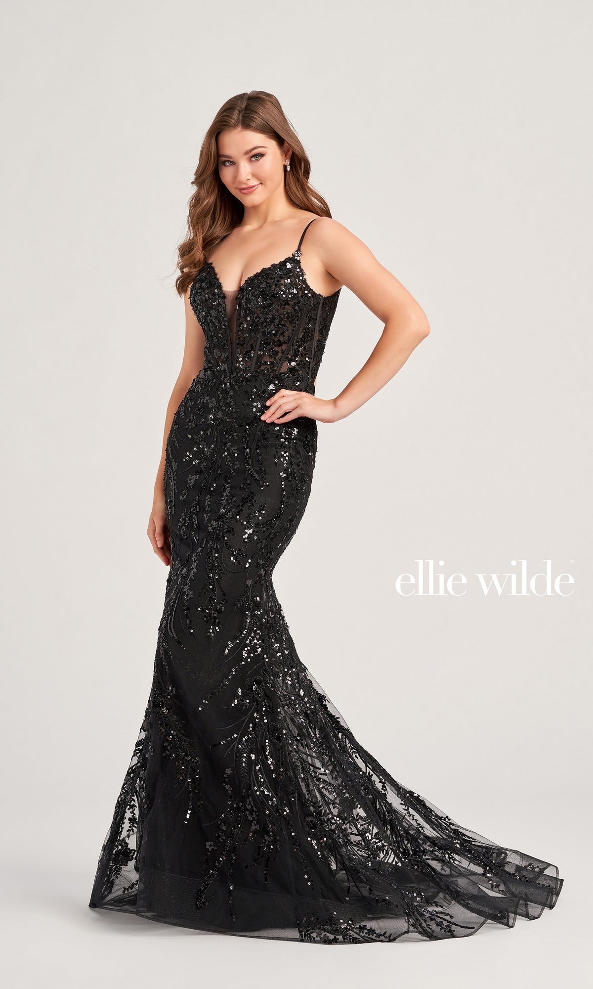 A woman poses in the Long Black Sequin Ellie Wilde Prom Dress EW35039, showcasing its fitted, floor-length mermaid silhouette with thin straps. The background is plain and light-colored, with